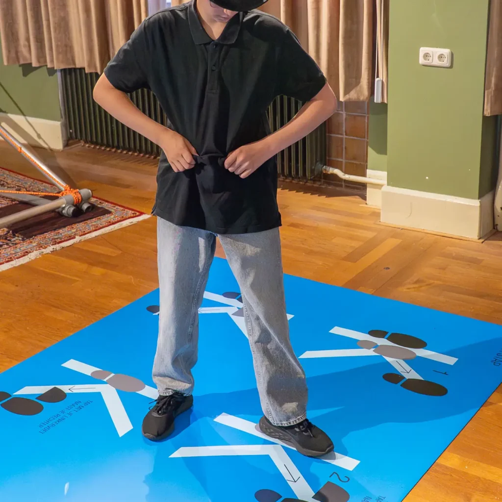 Een kind staat op een blauwe mat omringd door grote schoenveteronderdelen als onderdeel van een speelse activiteit in het Grand Hotel van Okke de Spin.