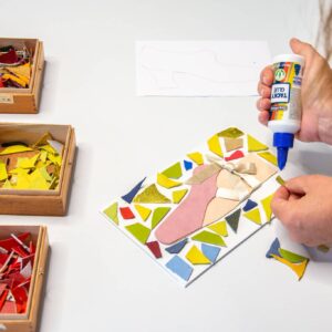 Een kind lijmt stukjes gekleurd leer op een wit paneeltje tijdens een mozaïekworkshop in het Schoenenkwartier.