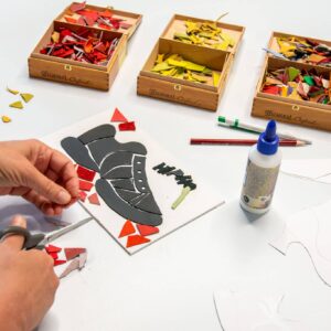 Een kind werkt aan een mozaïek van een schoen met zwarte en rode leerresten, naast een bakje met materialen en een lijmflesje.
