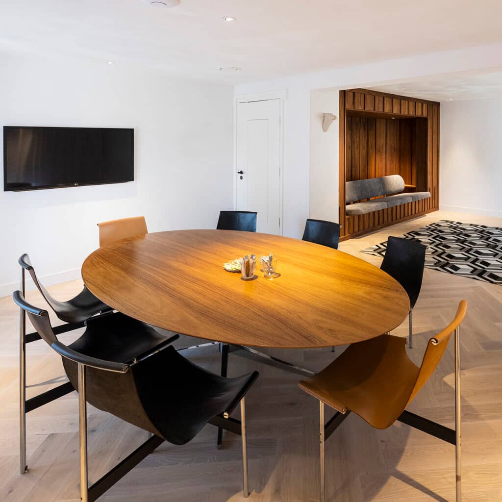 De Business Lounge van het Schoenenkwartier in Waalwijk: een lichte, stijlvolle vergaderruimte met een ovale houten tafel, leren stoelen, een flatscreen en een houten zitbank op de achtergrond.